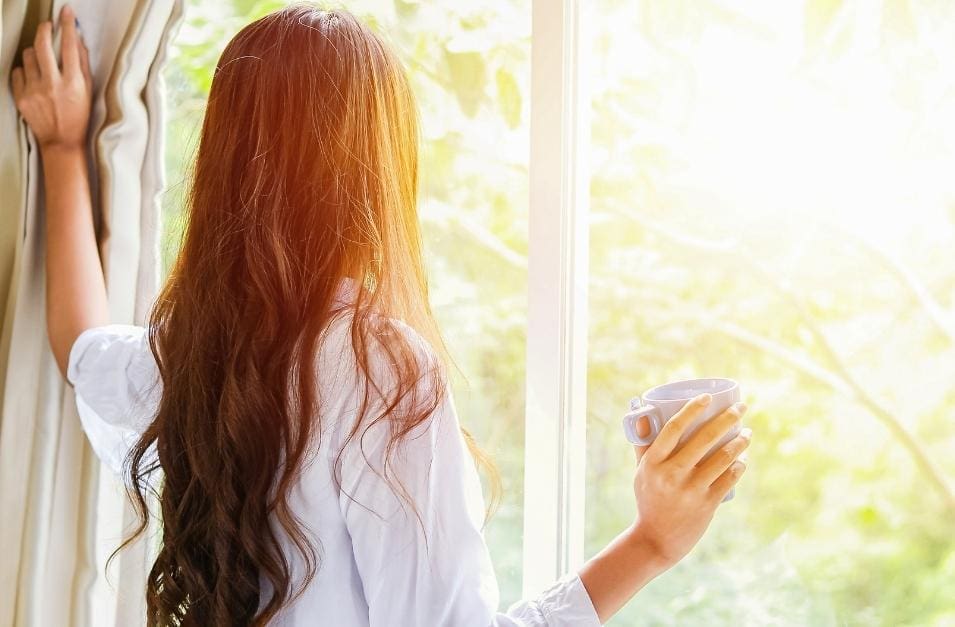 A woman, coping with loneliness when working alone, looks out the window while holding a cup of coffee.